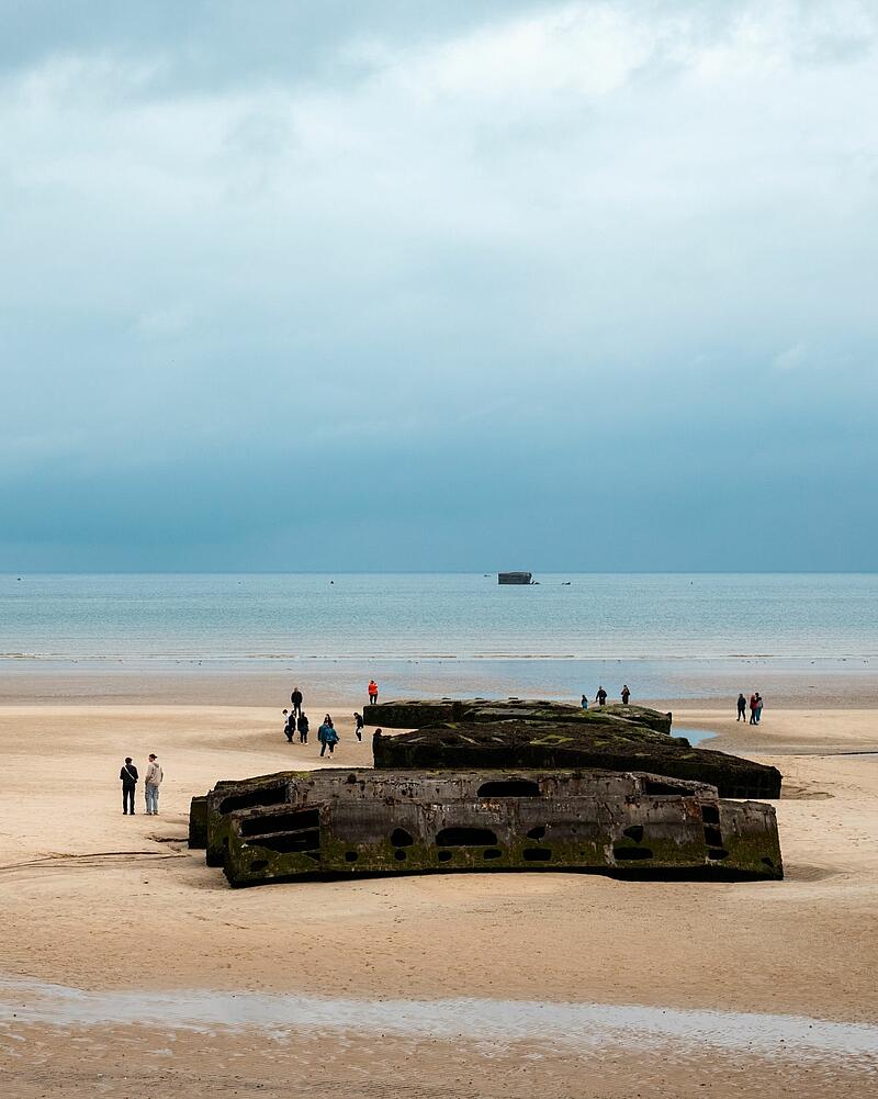 [Translate to Néerlandais:] Wide shot of a sandy beach under an overcast sky, where tourists walk around large concrete blocks, an excursion easily accessible from Les Castels campings in Normandy.