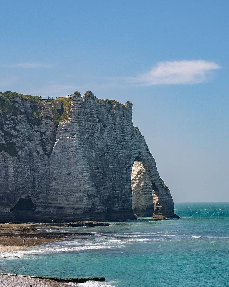 [Translate to Néerlandais:] A vertical shot of the famous towering white chalk cliffs and natural arch of Étretat, with waves crashing onto the beach, located near a Les Castels camping resort.