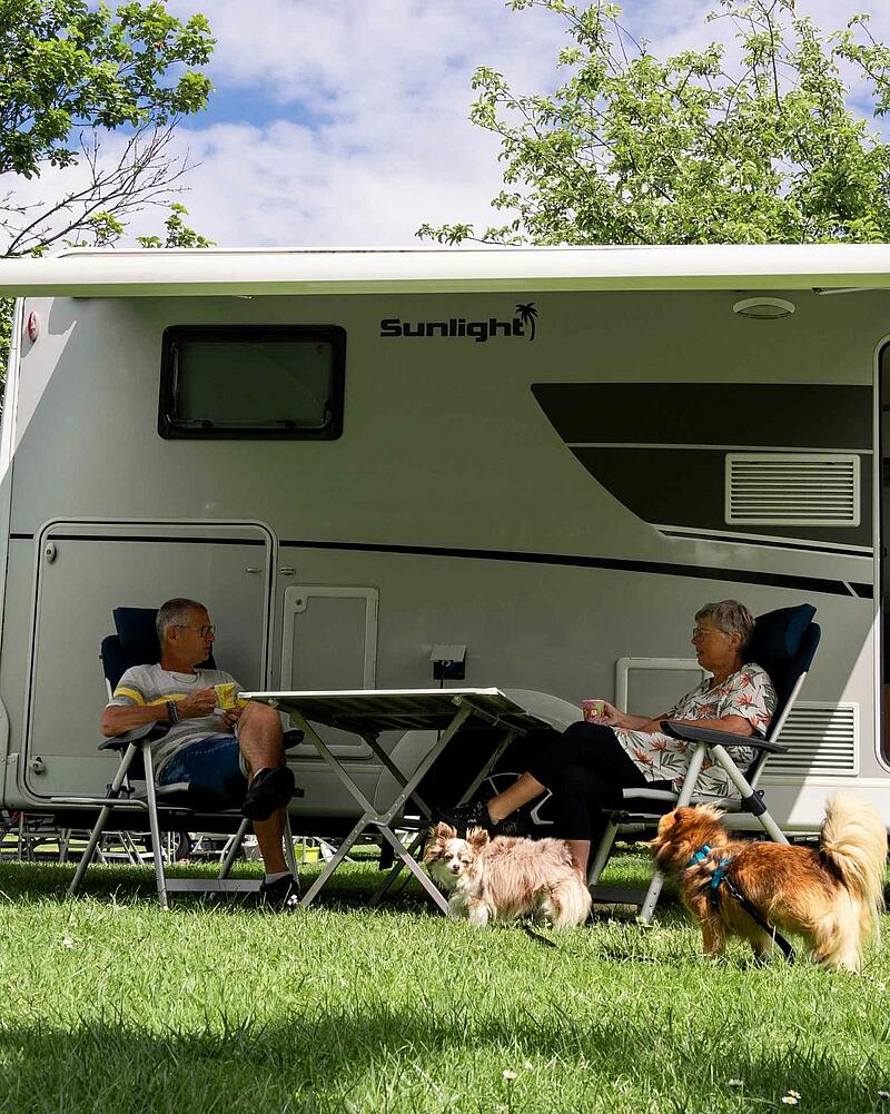 [Translate to Néerlandais:] A man and a woman sitting at a camping table under the awning of a modern Sunlight motorhome on a pet-friendly pitch at Les Castels.
