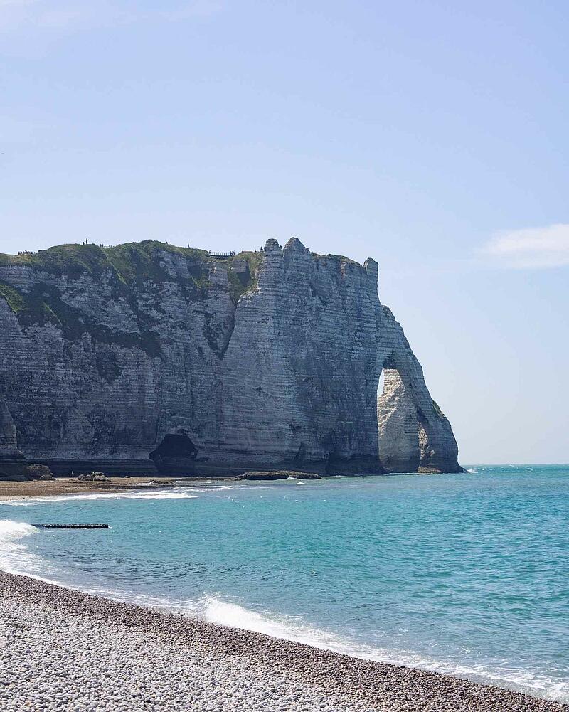 [Translate to Néerlandais:] The dramatic white chalk cliffs and natural rock arch of Étretat rising above a pebble beach and the turquoise waters of the sea in Normandy.