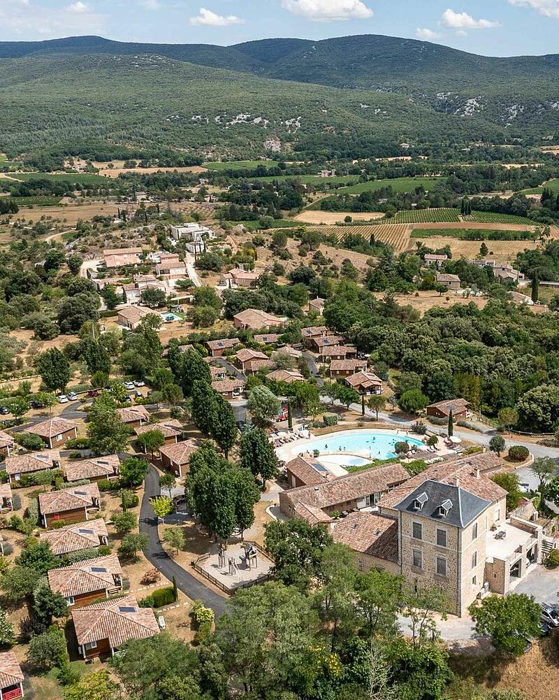 [Translate to Néerlandais:] Drone perspective of the heated outdoor swimming pool, stone estate building, and eco-friendly wooden chalets surrounded by nature at a 5-star Les Castels campsite.