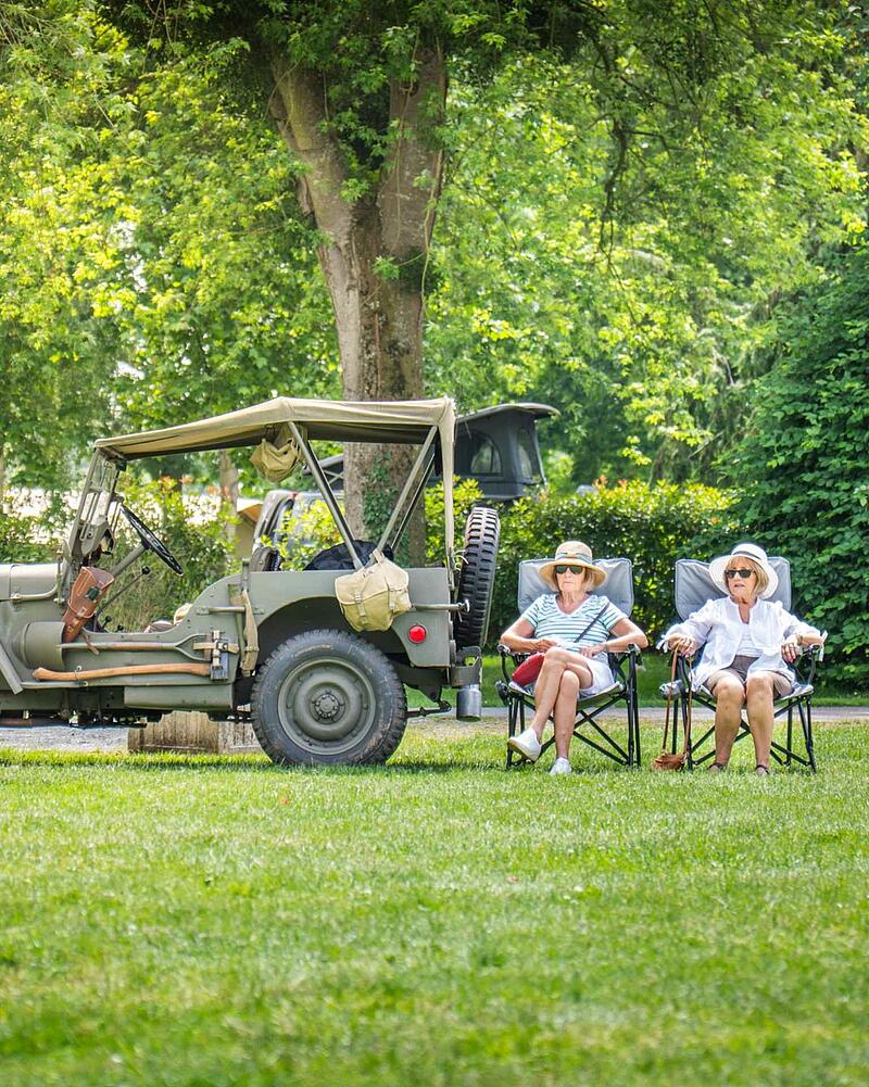 [Translate to Néerlandais:] Two senior women wearing sun hats sitting on camping chairs on a green lawn next to a classic green vintage military jeep at a Les Castels campsite.