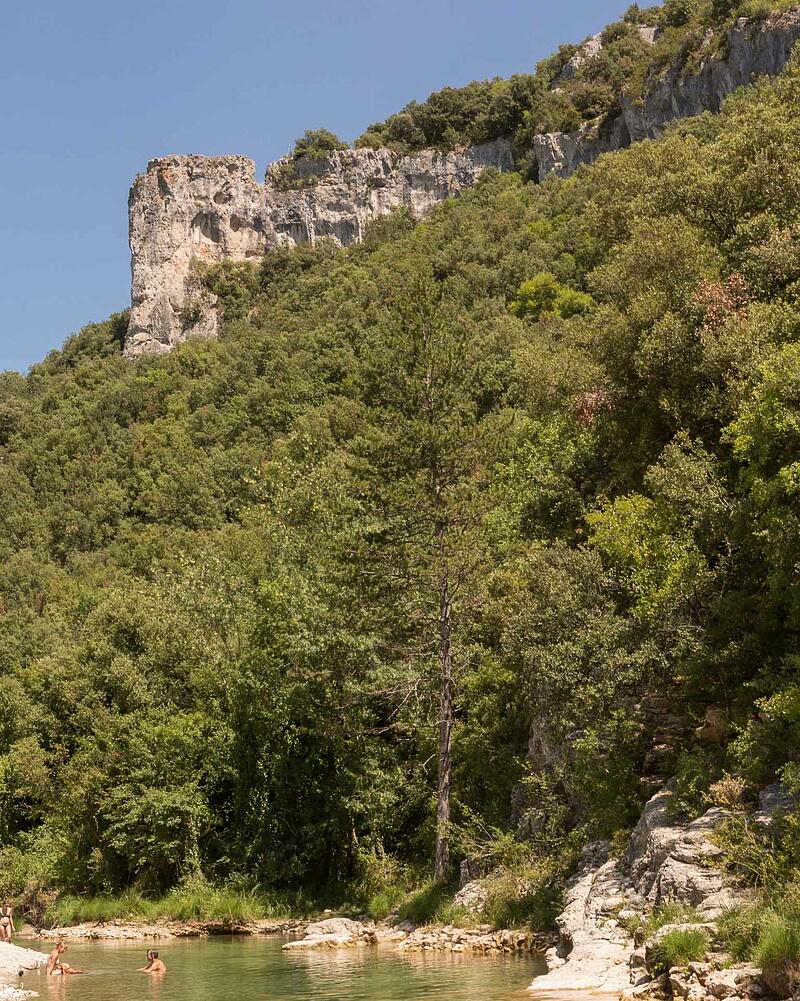 [Translate to Néerlandais:] People swimming and relaxing in the clear river water at the foot of towering limestone cliffs in the Ardeche Gorges near a luxury Les Castels estate.