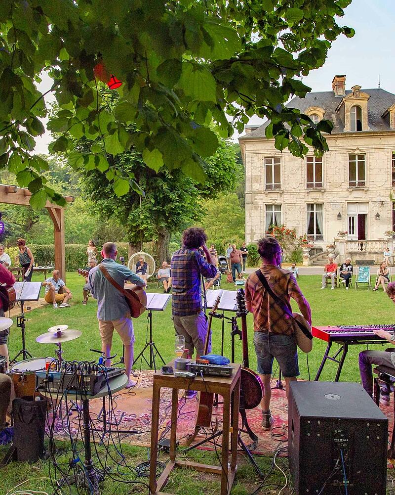 [Translate to Néerlandais:] Stage view from behind a live band playing guitars and keyboards outdoors for an audience seated on the grass during a premium evening event at Les Castels.