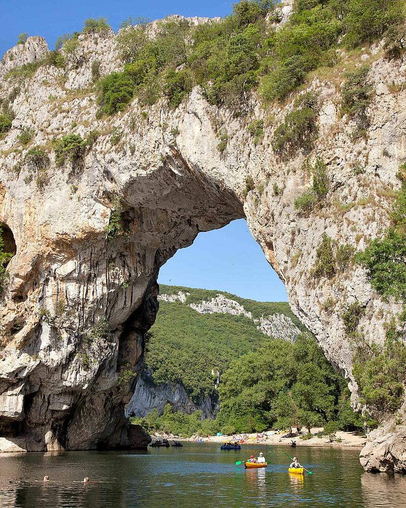 [Translate to Néerlandais:] Kayaks and canoes floating on the river under the massive, natural limestone arch of the Pont d'Arc in the spectacular Ardèche Gorges.