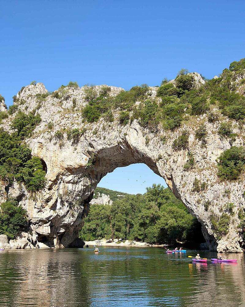[Translate to Néerlandais:] Colorful holidaymakers paddling on kayaks and canoes on the river right underneath the giant natural stone bridge of Pont d Arc near Les Castels luxury campsites.