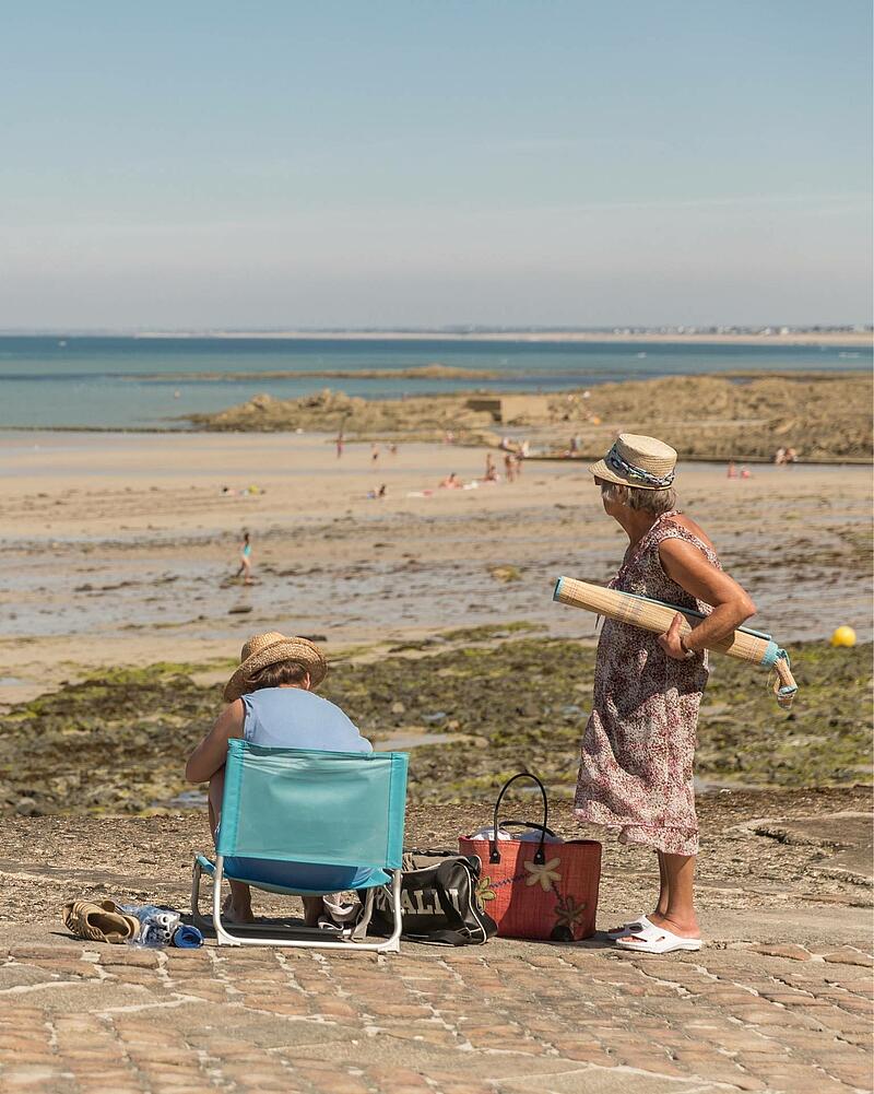 [Translate to Néerlandais:] Two women wearing straw hats spending a sunny day on a stone seawall, overlooking a vast sandy beach during a seaside holiday with Les Castels campings.