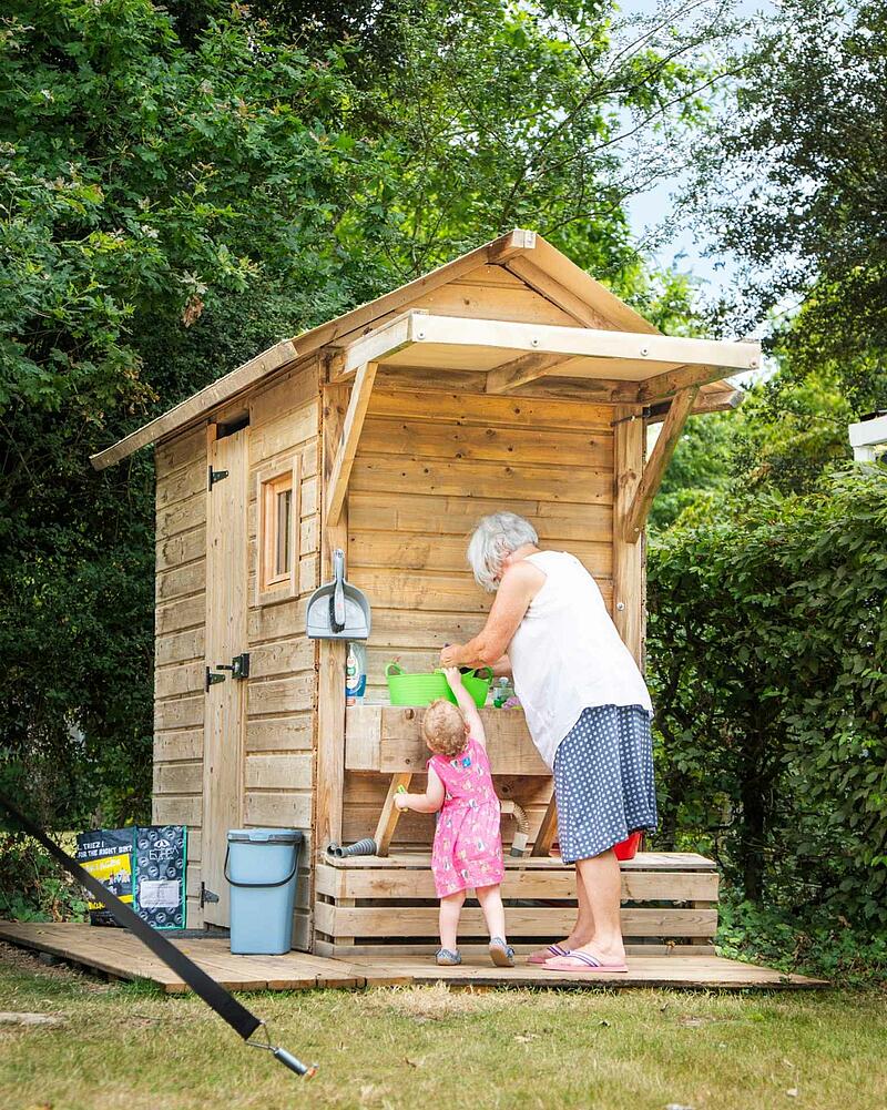 [Translate to Néerlandais:] Woman and young girl interact at a rustic wooden outdoor kitchen station on a Les Castels camping estate.