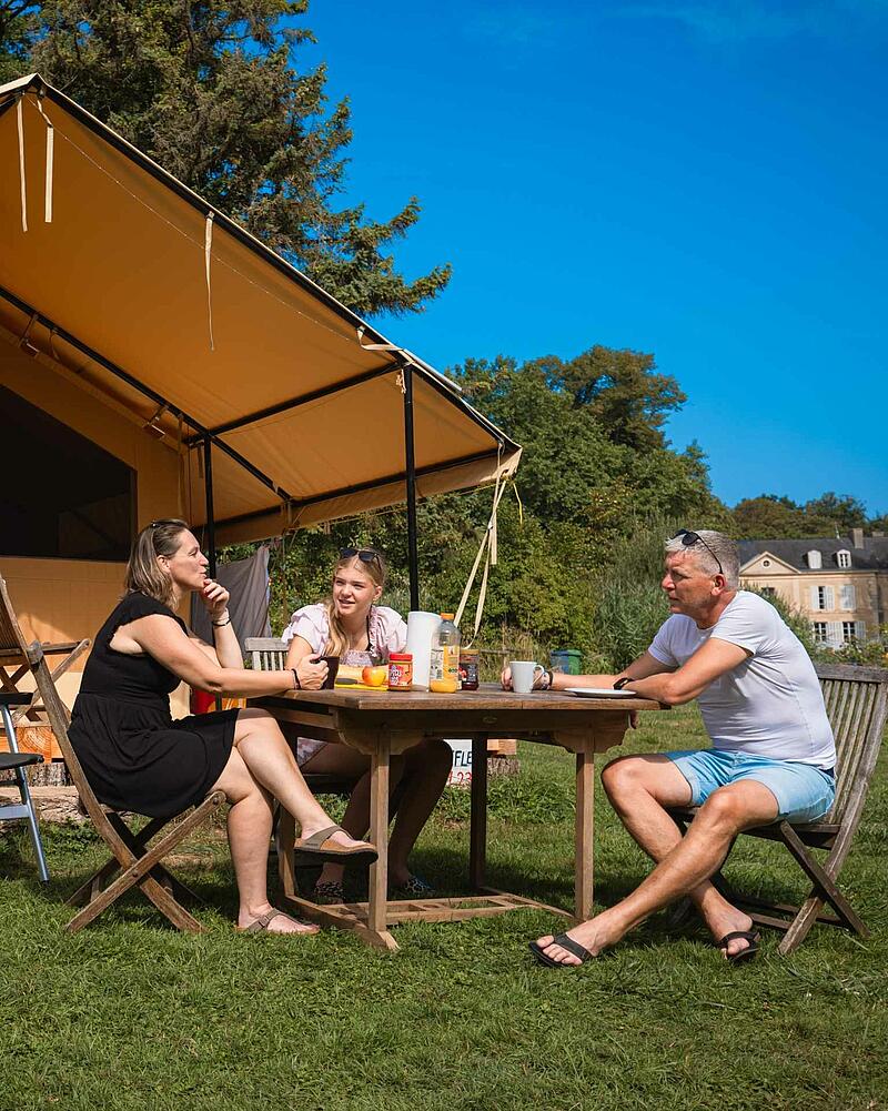 [Translate to Néerlandais:] A mother, father, and daughter sit around a wooden picnic table having breakfast outside a large luxury canvas glamping safari tent on a sunny campsite lawn.