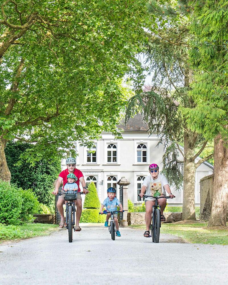 [Translate to Néerlandais:] A family of four, including two young children wearing helmets, cycling towards the camera on a gravel path during their holiday at a Les Castels domain.