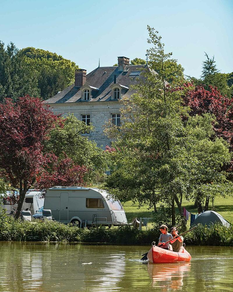 [Translate to Néerlandais:] An orange canoe with a man and a young boy paddling on a calm lake, with a caravan parked on the grassy bank of a Les Castels estate.
