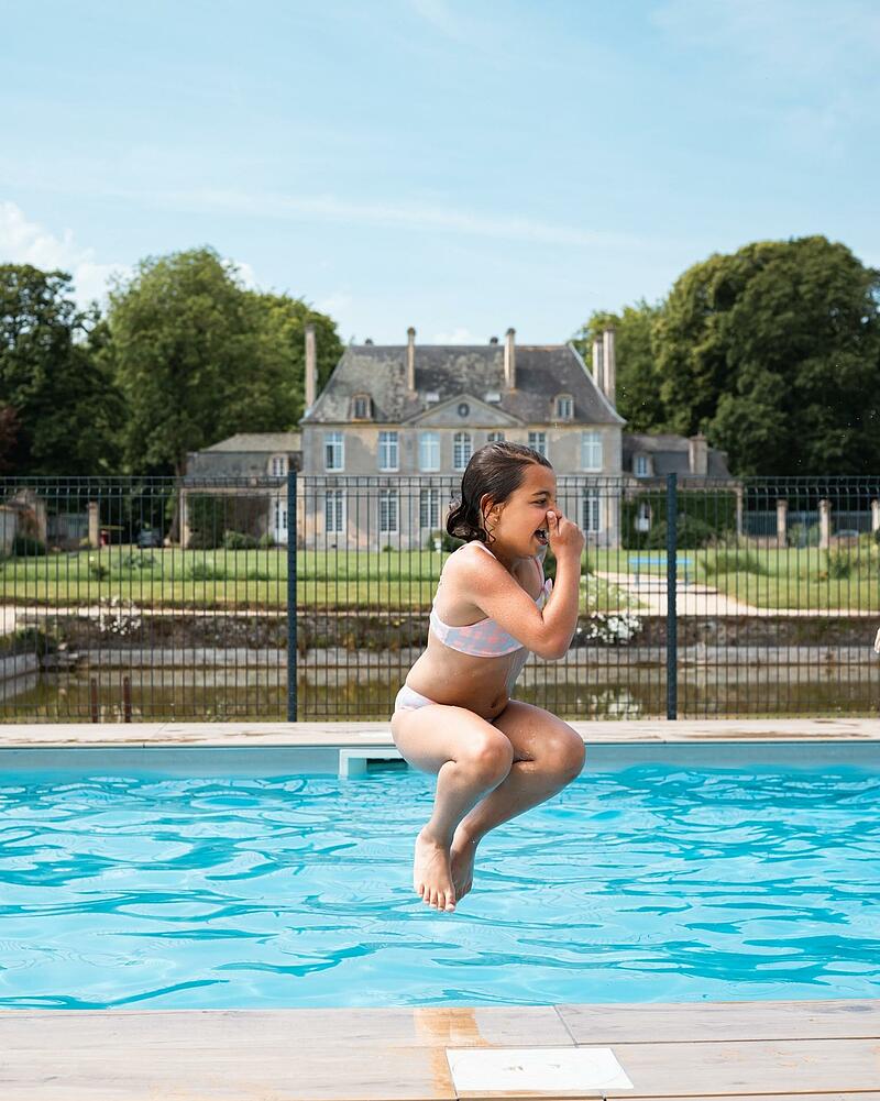 [Translate to Néerlandais:] A young girl mid-air in a cannonball jump into a crystal-clear outdoor swimming pool, located in the gardens of a grand French estate at Les Castels.
