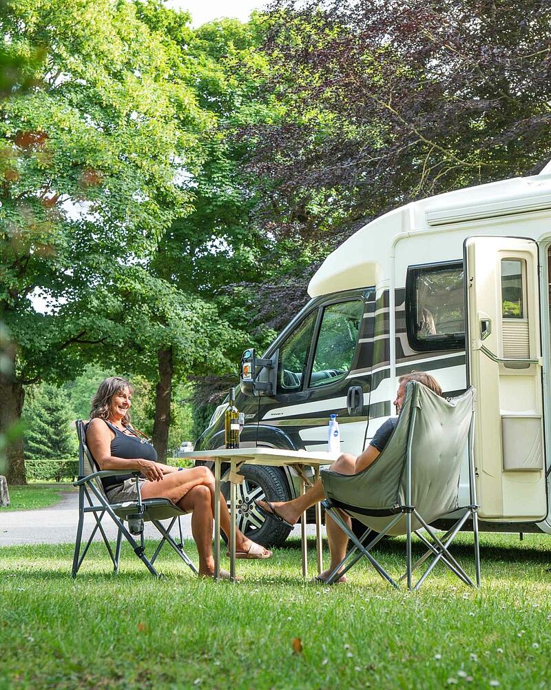 [Translate to Néerlandais:] A man and a woman sit in comfortable folding chairs at a camping table with a bottle of wine, relaxing next to their modern motorhome on a shaded, grassy pitch.