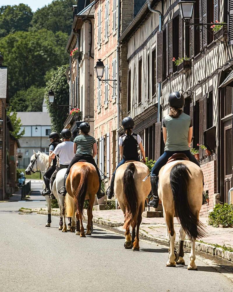 [Translate to Néerlandais:] A group of riders on horseback walking down a village street lined with classic half-timbered stone houses, an outdoor activity proposed near your Les Castels camping resort.