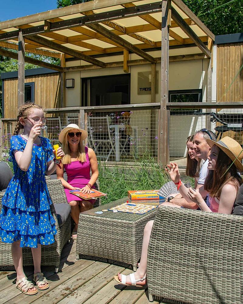 [Translate to Néerlandais:] A family enjoys quality time together playing a board game and blowing bubbles on the spacious wooden terrace of a premium, modern mobile home at a Les Castels campsite.
