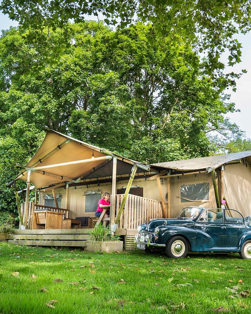 [Translate to Néerlandais:] A large canvas glamping safari tent with a wooden porch set amongst lush green trees on a premium pitch at a Les Castels 5-star campsite.