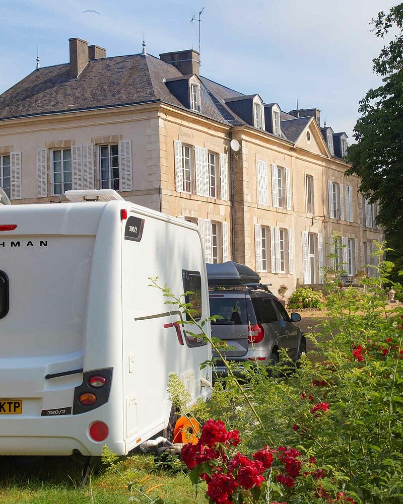 [Translate to Néerlandais:] The back of a white caravan and a grey SUV parked on a sunny grass pitch in front of a grand, historic stone chateau, beautifully framed by blooming red roses in the foreground.