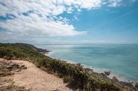 [Translate to Néerlandais:] A scenic coastal view in Normandy featuring a dirt pathway on a grassy cliff overlooking a vast expanse of turquoise sea under a blue sky with fluffy white clouds, perfect for an outdoor stay at Les Castels.